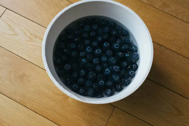 April 30, 2020 - Warsaw, Poland: blueberries being washed in large bowl of water on a wooden background - high above view