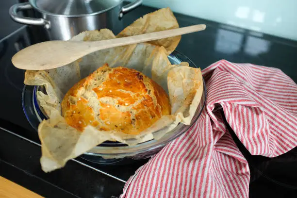 April 29, 2020 - Warsaw, Poland: homemade sourdough white bread loaf in a glass pan and wax paper - tasty, orange crust, wooden spoon, linen dish towel, domestic kitchen stove top
