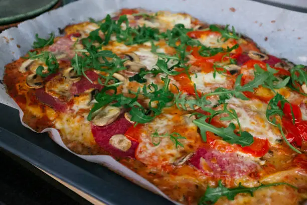 May 1, 2020 - Warsaw, Poland: close-up of a homemade and handmade pizza on a baking sheet with salami, arugula, paprika, mozarella cheese and freshly made tomato sauce in a domestic kitchen, real life.
