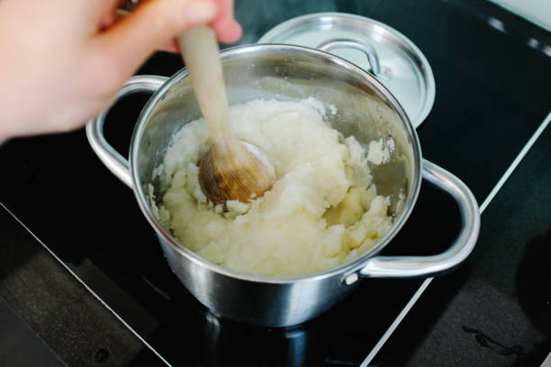 istock Preparing mashed potatoes in a domestic kitchen - woman's hand mashing potatoes with wooden masher in a metallic cooking pot with lid nearby on stove top. 1222773381