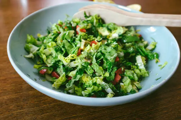 April 30, 2020 - Warsaw, Poland: large bowl of on green salad full of vitamins, green raw vegetables - lettuce, avocado, paprika in navy blue plate on a wooden kitchen counter in domestic kitchen