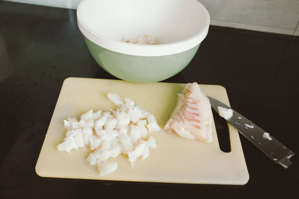 Preparing raw cod fish on a white plastic cutting board in domestic kitchen - chopped fish pieces for stew, high angle, close up view, healthy eating, vegan food stock photo