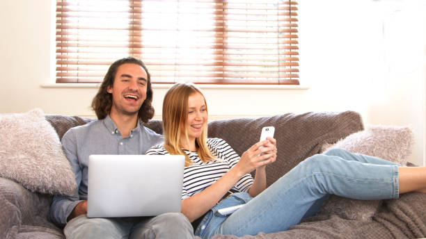 Young couple watching tv and using wireless technology. A young hetrosexual couple in thier 20s sitting on a sofa at home. They are watching tv, while the man has a laptop the young woman is holding her smartphone with her feet up. Dominic stock pictures, royalty-free photos & images