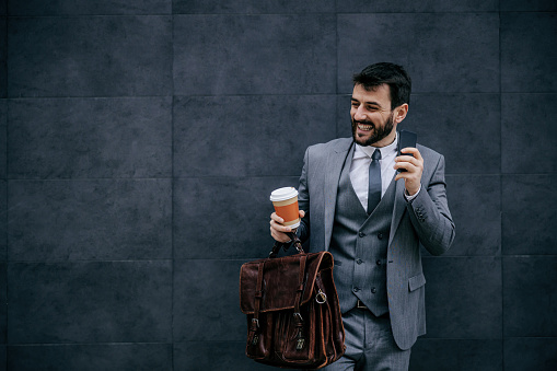 Young smiling fashionable businessman leaning on the wall and holding smart phone.