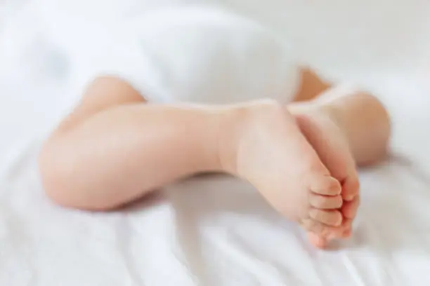 Baby's feet. Bare heels of little child wearing white bodysuit and diaper. Cozy morning bedtime at home.
