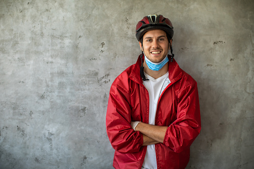 Portrait of delivery person during coronavirus pandemic, wearing red jacket, standing near gray wall with arms crossed, looking at camera and smiling