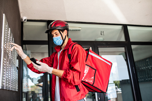 Delivery man wearing red jacket, cycling helmet, protective face mask and surgical gloves, checking the right address using smart phone while standing in front of intercom during corona virus pandemic