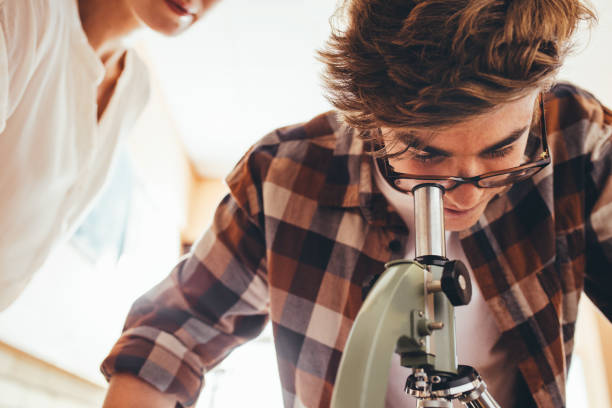 Student looking through a microscope in class Boy with female teacher in school laboratory looking in a microscope. Male student looking at slides through a microscope in class. biology class stock pictures, royalty-free photos & images