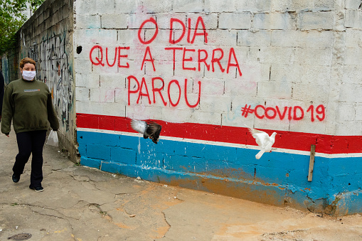 Sao Paulo, Brazil - April 17, 2020: Woman wearing protective face mask, walking by a street wall with graffiti about the Pandemic with “#Covid 19,” as birds fly by.
