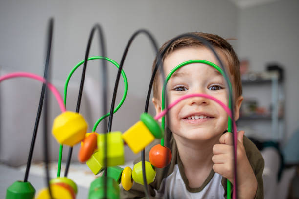 An adorable happy toddler boy sits at a home in a living room and reaches up cheerfully to play with a toy bead maze. Happy toddler boy playing with toys at home. Toddler play with a color educational toy