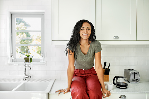 Portrait of smiling woman sitting on kitchen counter. Happy young female is relaxing at home. She is wearing casuals during lockdown.