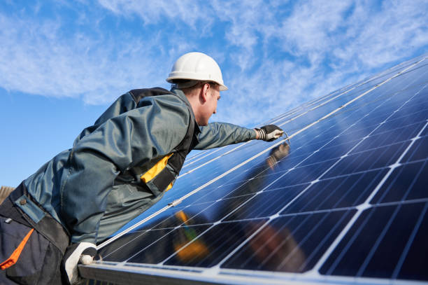 Workman standing on the ground trying to fasten solar batteries together, installing process Side view snapshot of workman, wearing uniform, working gloves and helmet, setting a shiny new solar battery with help of hex key, blue sky on background. Green energy concept solar power station photos stock pictures, royalty-free photos & images