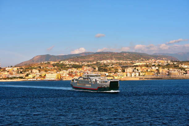 Ferry sailing in the Strait of Messina between Sicily and Calabria. On the background the sicilian coastline and the city of Messina. Italy.