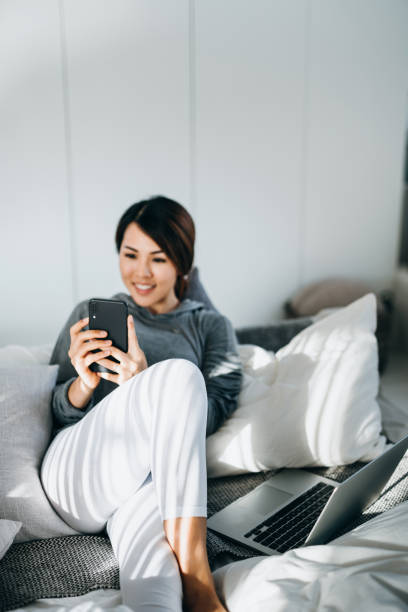 istock Smiling young Asian woman using smartphone while lying in bed, with laptop by the side 1222815294