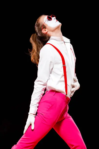 Photo of Portrait of male mime artist, isolated on black background. Man in suspenders and pink trousers is posing touching his stomach. Symbol of popular, dance, 90s