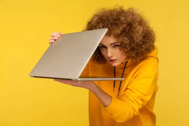 Woman with curly hair in urban style hoody peeking out half-closed laptop with sly suspicious look, spying and knowing secret information, using internet service without permission. isolated on yellow