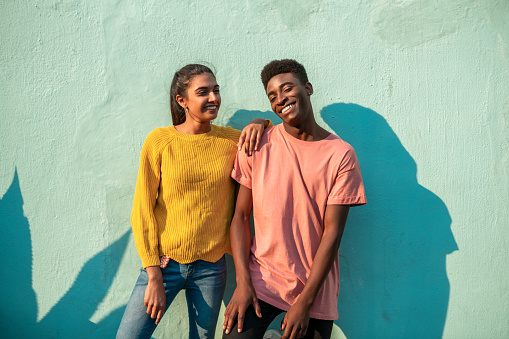 Portrait of Young happy couple. They are looking at camera while leaning on light blue wall.
