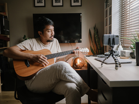 Hipster asian man learns to play the guitar with the help of an online tutorial at home.