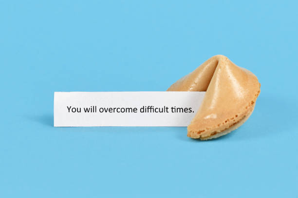 Fortune cookie with motivational text on paper saying 'You will overcome difficult times' on blue background Whole unopened fortune cookie with motivational text on white paper saying 'You will overcome difficult times' on blue background english spoken stock pictures, royalty-free photos & images