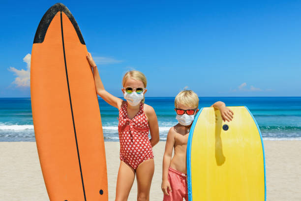Young surfers wearing sunglasses, protective mask on sea beach Funny kids with surf boards wear protective mask on sea beach. Cancelled cruises, tours due coronavirus COVID 19 world epidemic. Travel ban for family vacation, tourism industry crisis at summer 2020 raro stock pictures, royalty-free photos & images