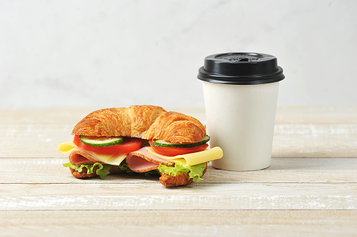 Sandwich with cheese and ham and a paper cup with coffee. Takeaway food concept. Light wooden background. Close-up.