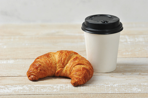 Croissant and a cup of coffee. Takeaway coffee and takeaway concept. Wooden light background.
