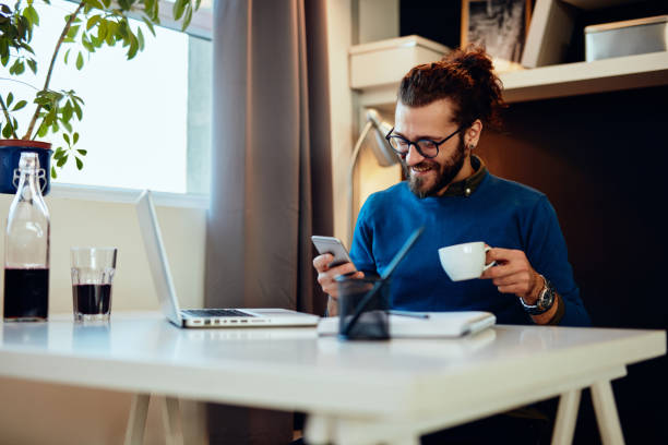 Attractive caucasian bearded entrepreneur sitting in his office, using smart phone and taking a break from work.