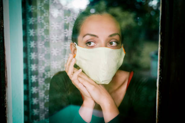 istock Young woman in quarantine wearing a mask and looking through the window 1222834708