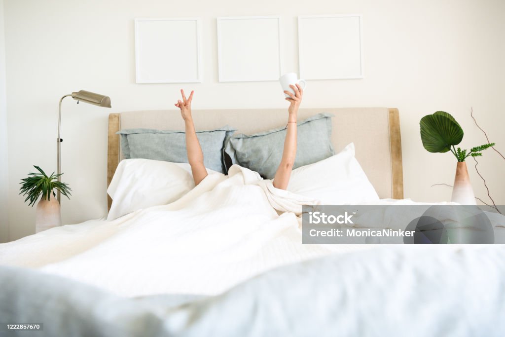 Hispanic mature woman waking up in bed with coffee mug in hand Hispanic mature woman waking up in bed with coffee mug in hand underneath the bed cover Bed - Furniture Stock Photo