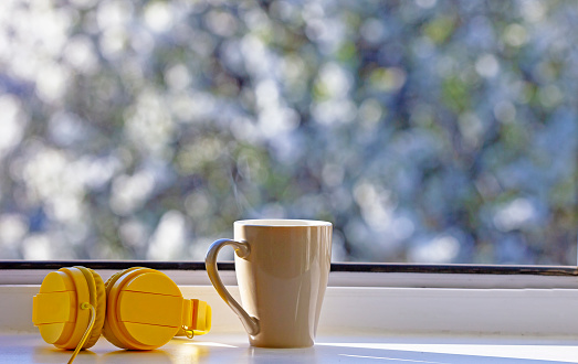 cup of hot tea with headphones on a window sill near an open window in the morning, followed by cherry blossoms, quarantine, stay home, horizontal format