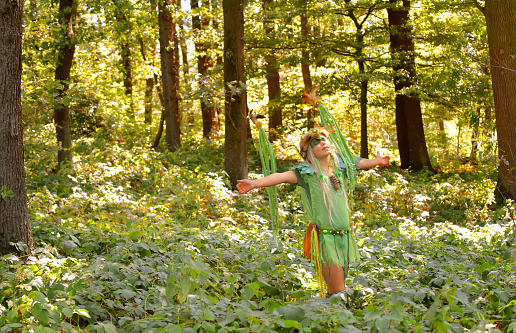 A young girl is dressed up as a fairy.\nShe is seen in a forest with a large\ndecorated wreath on her head.