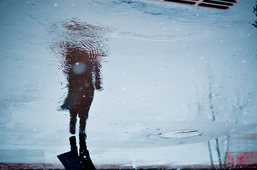 Depressed and loneliness mood concept. Blurry reflection in a rain puddle of alone walking person on wet city street during rainy weather.