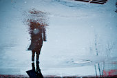 Blurry reflection in a rain puddle of alone walking person on wet city street.