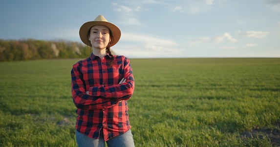 Portrait of smiling woman farmer in hat, red checkered shirt standing in a field with green wheat with crossed her arms on sunset