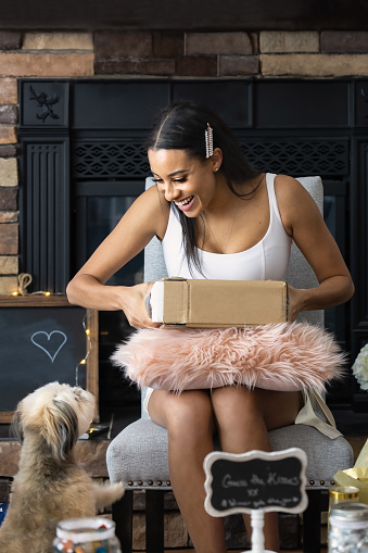 Young smiling woman opening a present in her home while looking at her small fluffy puppy