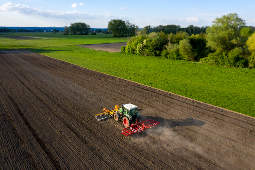 Tractor sowing seed on plowed field. Sowing seeds of corn and sunflower. Blue Tractor with disk harrow on plowing field. Seeding machinery on farm field. Seed sowing in farmland, aerial view.