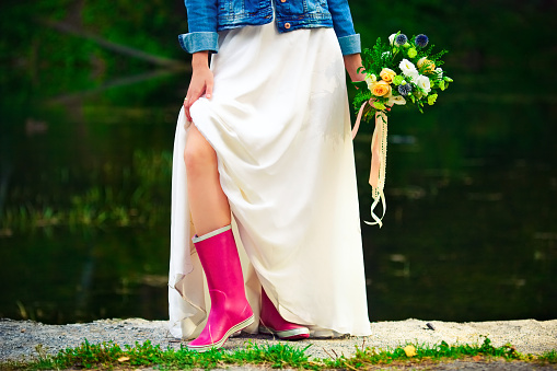 bride in white dress and pink rubber boots is holding a bouquet of flowers