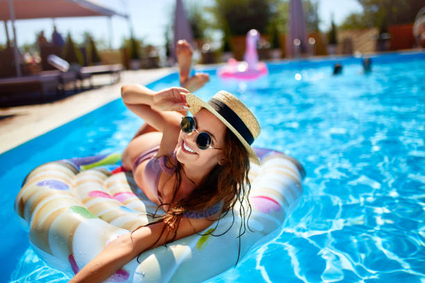Young smiling fitted girl in bikini, straw hat relax on inflatable swan in swimming pool. Attractive woman in swimwear lies in the sun on tropical vacation. Pretty female sunbathing at luxury resort stock photo