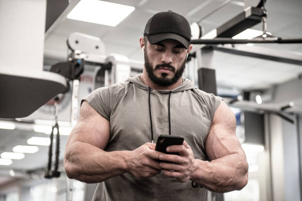 homem forte jovem barbudo com relógio boné no smartphone celular durante a pausa de treino de treino - strongman - fotografias e filmes do acervo