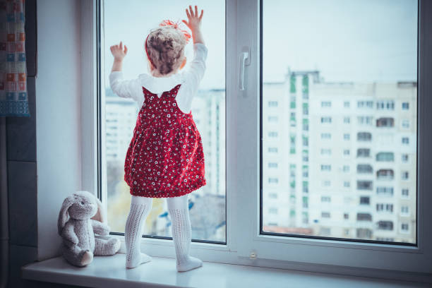 dziewczynka maluch stoi na parapecie, patrząc przez okno na wysokich budynkach i miękkie zabawki królik obok niej - violence child abuse child baby girls zdjęcia i obrazy z banku zdjęć