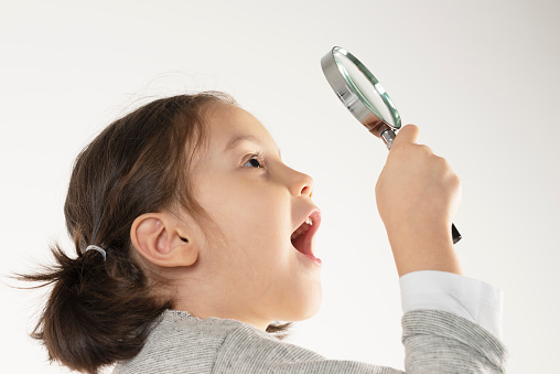 Side view of a caucasian little girl who is holding a magnifier glass in hand and is making a surprised happy gesture.