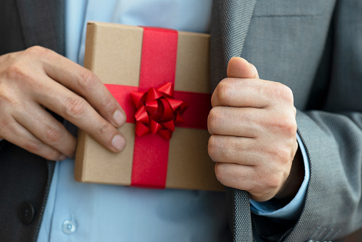 Chest view of an unrecognizable businessman wearing a suit who is about to give a gift box out of his jacket. The gift box is wrapped with red ribbon.