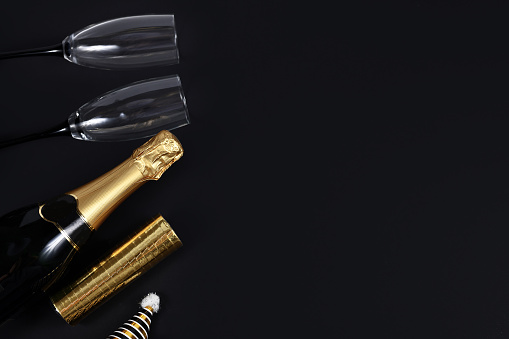 Champagne bottle head and drinking glasses, striped party hat and golden paper streamers on left side of dark black background with blank copy space.