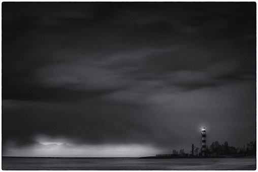 Dramatic cloudy sky over Baltic sea. Sea waves at long exposure. Striped lighthouse on the seashore. Framed black and white.
