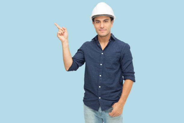 istock Handsome man industrial engineer wearing a white hard helmet and pointing finger towards something blank copy space isolated on light blue background 1222934194