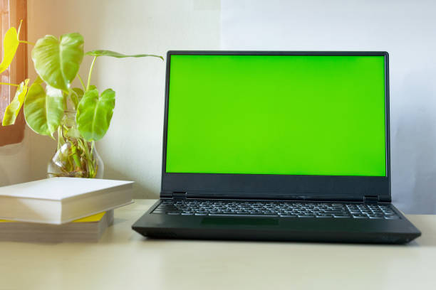Laptop computer showing chroma green screen on LCD display stands on a desk with books next to ready for further design and copy space. stock photo