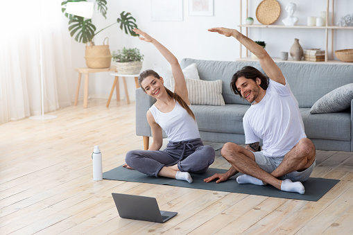 Healthy couple exercising at home, sitting on sport mat and watching videos on laptop, copy space