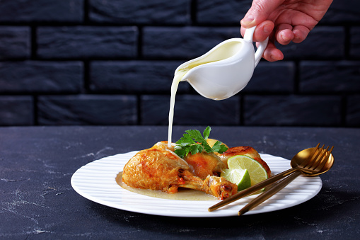 Woman hand is pouring dripping coconut sauce on top of roasted chicken leg served with baby potatoes on a white plate, horizontal view from above