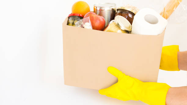 istock Courier, delivery man in yellow gloves holding craft box with different products, various canned food, pasta and cereals. 1222944450