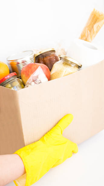 istock Courier, delivery man in yellow gloves holding craft box with different products, various canned food, pasta and cereals. 1222944487